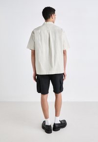Camisa de manga corta en color beige claro con un pliegue en la parte trasera, combinada con shorts cargo negros con bolsillos laterales y zapatos negros con suelas gruesas.