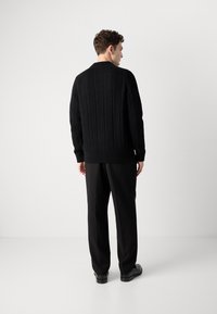 Μαύρο πλεκτό πουλόβερ με ρίγες, μακριά μανίκια, στρογγυλή λαιμόκοψη και χαλαρή εφαρμογή, σε συνδυασμό με μαύρο παντελόνι και παπούτσια, όπως φαίνεται από πίσω.