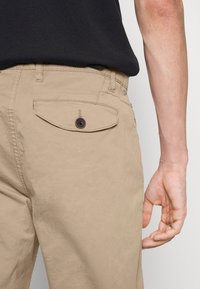 Beige Baumwollhose mit einer Gesäßtasche, die über einen Klappverschluss mit braunem Knopf verfügt. Glatte Textur, gerader Schnitt und mittelhoher Bund.