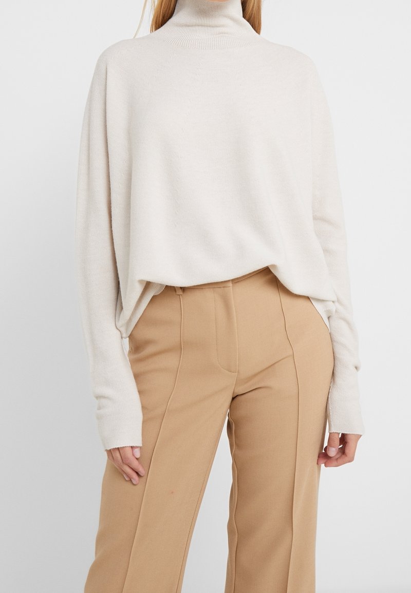 Pull à col roulé crème avec une coupe décontractée, des manches larges et une texture côtelée, associé à un pantalon fluide beige taille haute.