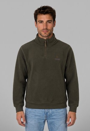 Hombre con forro polar verde oscuro con cremallera hasta el pecho y vaqueros azul claro, de pie frente a un fondo gris liso.