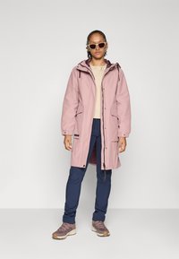 Lys rosa vanntett parka med hette, to frontlommer og justerbare mansjetter, kombinert med marineblå slim-fit bukser og gråbrune turstøvler.