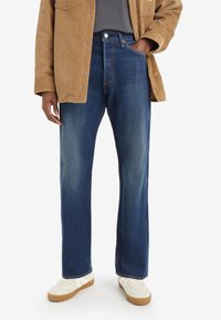 Jeans in denim blu scuro con taglio dritto, caratterizzati da cuciture a contrasto e tasche frontali. Indossati con una giacca in corduroy beige.