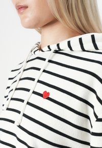Gros plan sur une personne portant un hoodie blanc avec des rayures noires et un petit cœur rouge brodé sur la poitrine, montrant la mâchoire et le cou.