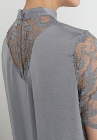 Blusa gris con un cuello alto, que presenta mangas de encaje transparente y un cierre de botones en la parte posterior. La tela es suave y de textura lisa.