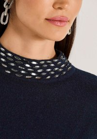 Nahaufnahme des unteren Gesichts einer Frau mit rosa Lippen, die einen dunkelblauen Pullover mit silbernen ovalen Verzierungen am Kragen und Strassohrringen trägt.