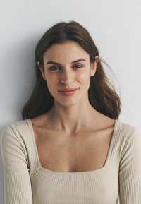 Eine Person mit langen braunen Haaren trägt ein hellbeiges, geripptes Oberteil mit quadratischem Ausschnitt, das feine Nähte entlang der Kanten aufweist.