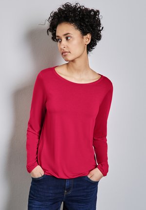 Chemise à manches longues de couleur rouge vif, en tissu doux, avec un col rond et des poignets élastiques, assortie à un jeans bleu foncé.