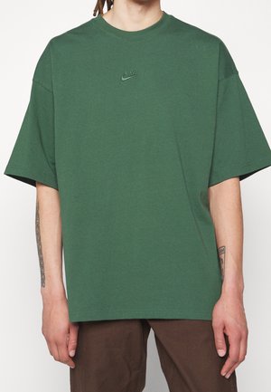 Osoba nosząca luźną, ciemnozieloną koszulkę Nike z krótkimi rękawami i małym wyszywanym logo, w połączeniu z brązowymi spodniami.