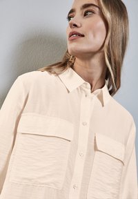 Camisa de botones color beige claro, hecha de una tela texturizada, con dos bolsillos en el pecho y un cuello clásico. Acabado suave, corte relajado.