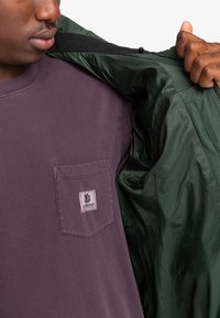 Giacca verde leggera aperta sopra una maglia viola scuro con taschino sul petto. La giacca presenta una texture liscia e lucida senza hardware visibili.