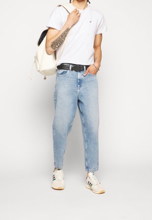 Personne portant un t-shirt blanc, un jean bleu clair, des baskets blanches rayées, portant un sac à dos blanc sur une épaule, debout devant un fond uni.