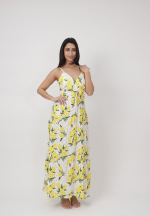 Femme debout, portant une longue robe blanche sans manches ornée de motifs de citrons jaunes et de feuilles vertes, posant devant un fond uni clair.