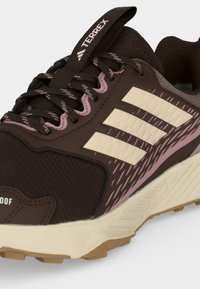 Καφέ και ροζ αθλητικό παπούτσι Adidas Terrex με κρεμ τρεις ρίγες, υφασμένο πέλμα και αδιάβροχη ετικέτα στην περιοχή του δακτύλου.