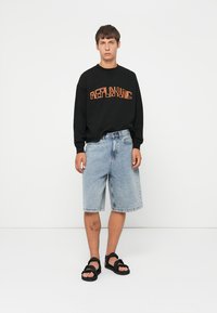Giovane uomo che indossa una maglietta nera a maniche lunghe con la scritta arancione "REPLAYING", pantaloni corti di denim chiaro fino al ginocchio, sandali neri, in piedi su uno sfondo bianco.