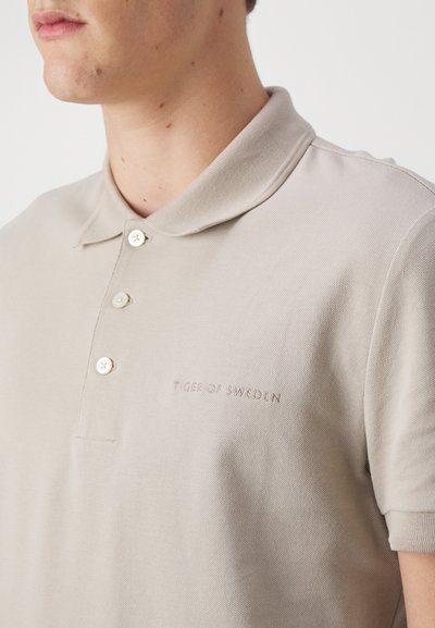 Beeži polo särk tekstuuriga kangast, millel on kolmest nupust koosnev kaelus ja rinnal tikitud "Tiger of Sweden" logo.