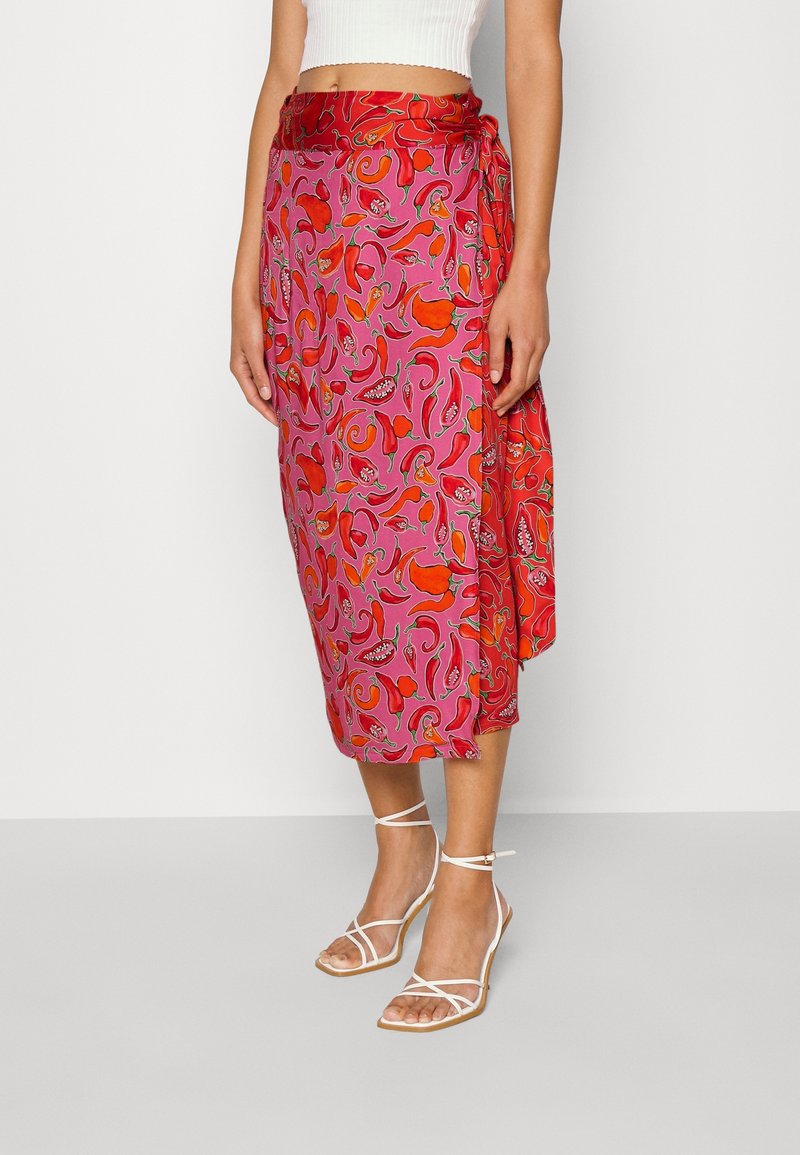 Femme portant une jupe midi portefeuille fleurie rose et rouge, un top court blanc et des sandales blanches à talons hauts à brides, debout sur un sol uni.