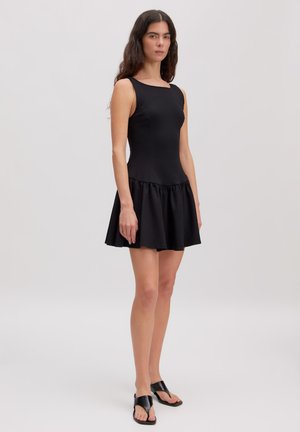 Woman with long dark hair wearing a sleeveless black dress with a flared skirt and black open-toe flat sandals standing on a plain background.