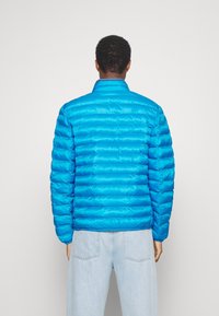 Giacca a vento blu con pattern a righe orizzontali, colletto alto e polsini elastici. Indossata sopra jeans in denim azzurro su uno sfondo piatto.