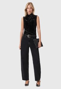 Blonde vrouw in een mouwloos zwart fluwelen top, hooggesneden zwarte jeans met riem, die een zwarte clutch vasthoudt en metallic puntige hakken draagt.