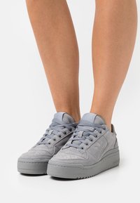 Gray suede sneakers with a textured finish, round toe, flat laces, and a thick rubber sole. Features logo on the side and tongue.