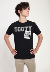 Černé bavlněné tričko s nápisem "SOOTY" ve starobylém bílém písmu a grafickým designem kapsy s kočkou v černobílé potisku.