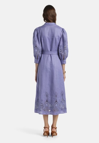 Frau trägt ein mittellanges lavendelfarbenes Kleid mit Puffärmeln und kunstvollen Spitzen-Einsätzen an Ärmeln und Saum, kombiniert mit braunen Sandalen mit hohen Absätzen.