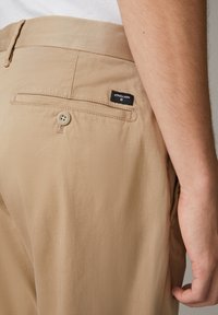 Beige Hose aus glattem Stoff, mit einer Gesäßtasche mit Knopfverschluss und einem kleinen schwarzen Etikett mit weißem Text.