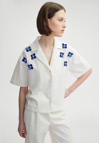 Camisa blanca de manga corta con adornos de flores de lentejuelas azules, cierre de botones en la parte delantera y un diseño de ajuste relajado. Tejido con textura sutil.