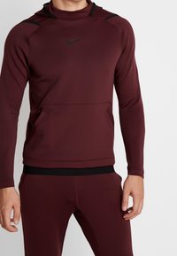 Bordeaux Nike hættetrøje med høj krave, sorte accentdetaljer, lange ærmer og en frontlomme. Glat, strækbart stof.