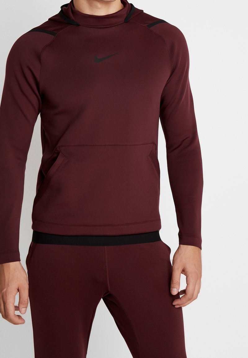 Bordeaux Nike hættetrøje med høj krave, sorte accentdetaljer, lange ærmer og en frontlomme. Glat, strækbart stof.