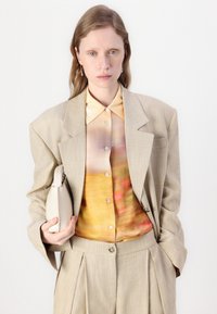 Μπεζ tailored κοστούμι με πατίνες στους ώμους, πουκάμισο με μοτίβο σε ζεστούς τόνους και μια μικρή ανοιχτόχρωμη τσάντα.