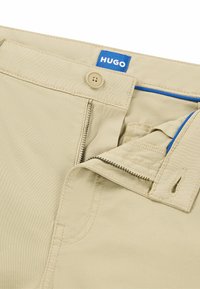 Khaki trousers with a blue HUGO tag, featuring a zipper fly, button closure, and contrasting blue stitching along the pocket edge.