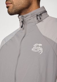 Pelēka viegla jaka ar rāvējslēdzēju un augstu apkakli, ar teksturētu audumu un izšūtu zīmolu uz krūtīm. Regulējama šņore apkaklē.