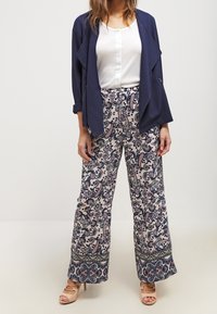 Blazer marine sur un haut blanc, associé à un pantalon large à motifs crème, marine et bordeaux. Des talons ouverts beiges complètent la tenue.