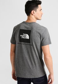 Camiseta gris de manga corta con un logotipo rectangular negro en la parte trasera, que dice "THE NORTH FACE" y "NEVER STOP EXPLORING."