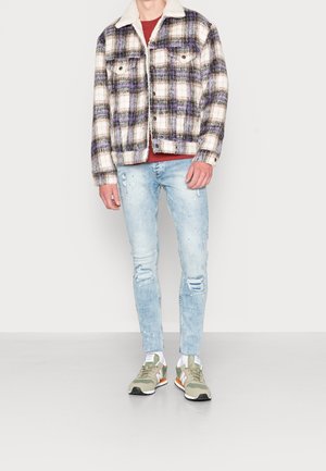 Homme portant une veste sherpa à carreaux violette, une chemise rouge, un jean skinny clair usé, des chaussettes blanches et des baskets beiges debout sur un sol gris.