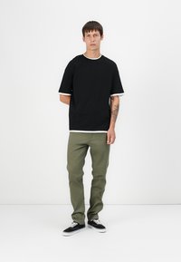 Black cotton t-shirt with white trim, paired with olive green pants. Model wears black sneakers, standing against a plain background.