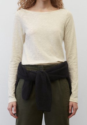 Vrouw draagt een lichtbeige top met lange mouwen, donkergroene broek en een zwarte gebreide trui gestrikt rond de taille.