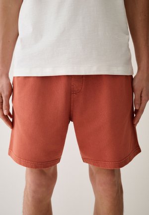 Uomo con pantaloncini arancioni bruciati e maglietta bianca, in piedi con le mani rilassate ai lati contro uno sfondo semplice.