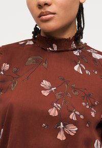 Gros plan sur une personne portant un chemisier couleur rouille avec des motifs floraux roses et un col montant à volants.