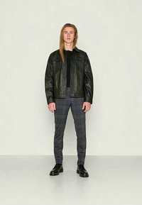 Casaco de pele preto, camisa preta, calças xadrez cinzas e sapatos pretos. Modelo em pé com as mãos nos bolsos num fundo claro.