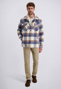 Kariertes Hemdjackett in Creme-, Blau- und Brauntönen, mit zwei Brusttaschen und einer durchgehenden Knopfleiste. Kombiniert mit beigen Hosen und braunen Schuhen.