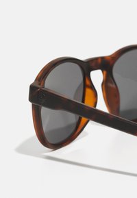 Óculos de sol em padrão tartaruga com lentes redondas e acessórios pretos. A armação apresenta um acabamento brilhante e hastes ligeiramente texturizadas.