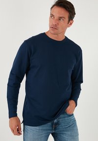 Camicia a maniche lunghe blu navy con scollo a giro, realizzata in tessuto morbido. Presenta una vestibilità rilassata e un orlo leggermente curvato, abbinata a jeans chiari.
