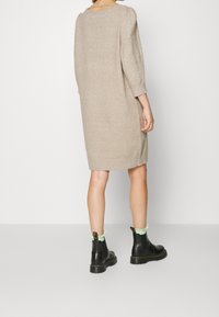 Beige stickad klänning med en avslappnad passform, ¾ ärmar och rund halsringning, matchad med svarta ankelstövlar med en grön accent.