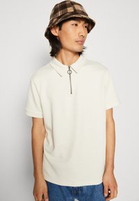 Cream-colored short-sleeve polo shirt with a textured diamond pattern, featuring a half-zip collar and a silver ring pull.