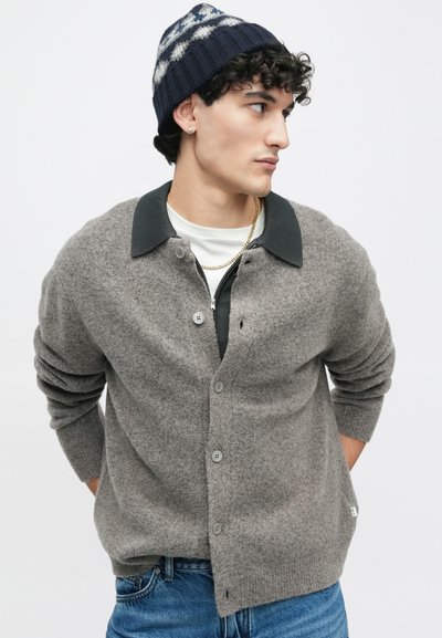 Szürke kötött kardigán sötétzöld gallérral, öt gombbal, bordás mandzsettával, fehér ing fölött viselve, kék farmernadrággal és mintás sapkával párosítva.