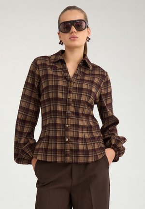 Femme portant de grandes lunettes de soleil foncées, une chemise à carreaux marron et beige boutonnée, et un pantalon brun foncé, debout les mains dans les poches.