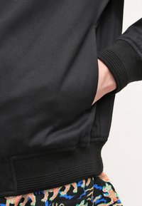Chaqueta bomber negra con una textura suave, puños y bajo acanalados. Detalles de bolsillo en los laterales, llevada sobre pantalones cortos con patrones que presentan diseños coloridos.